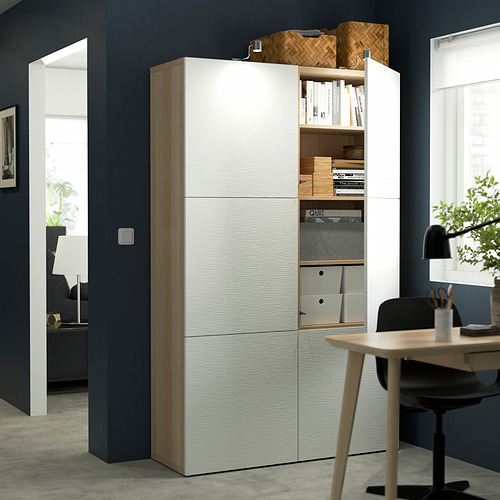 Un espacio de trabajo moderno con un mueble de almacenamiento BESTÅ blanco con puertas abiertas que muestran estantes con libros, cajas y otros artículos.