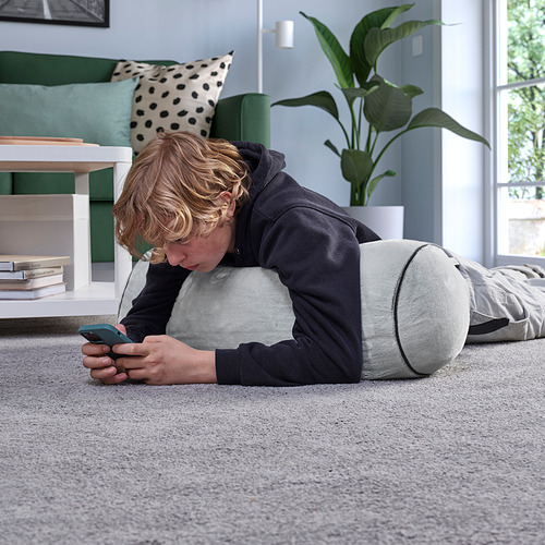 Niño acostado en un cojín BLÅSKATA gris, usando un teléfono inteligente en un piso alfombrado, con un sofá verde y una planta cerca.