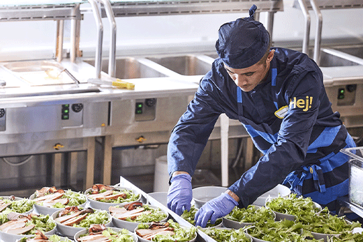 Un empleado del servicio de alimentos de IKEA con uniforme azul, gorro y guantes prepara cuidadosamente varias ensaladas de pollo en la línea de cafetería de una cocina comercial.
