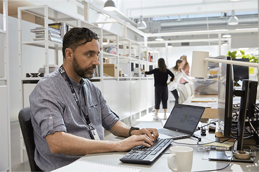 Un hombre con barba y un cordón de IKEA está sentado frente a un escritorio escribiendo en un teclado en una oficina moderna e iluminada.