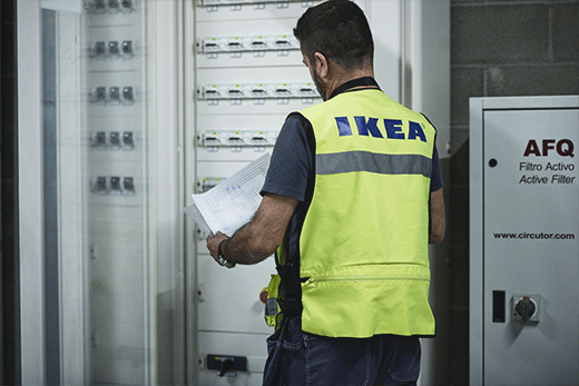 Una vista desde atrás de un trabajador de mantenimiento de IKEA con un chaleco de seguridad amarillo neón, sosteniendo documentos técnicos mientras inspecciona un gran panel eléctrico.