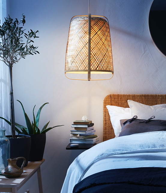 Bedroom with a lit hanging lamp made of natural fibers, woven headboard bed, decorative plants, and a stack of books on the bedside table.