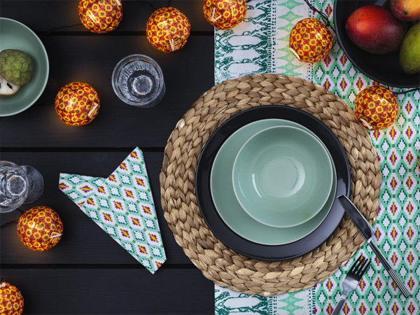 Table setting featuring green dinnerware, a woven placemat, and glowing string lights.