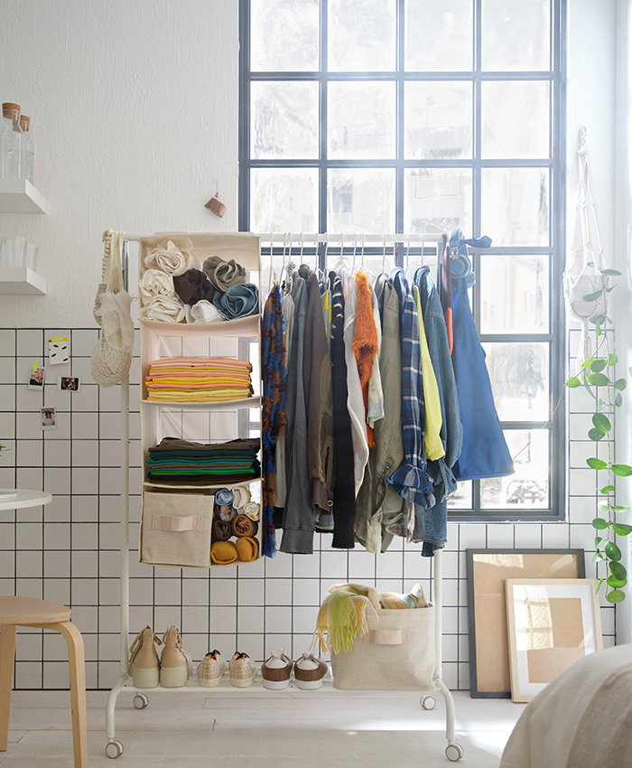 White clothing rack with hanging shelves and color-coordinated garments, placed by a large window and decorated with storage boxes, shoes, and plants.