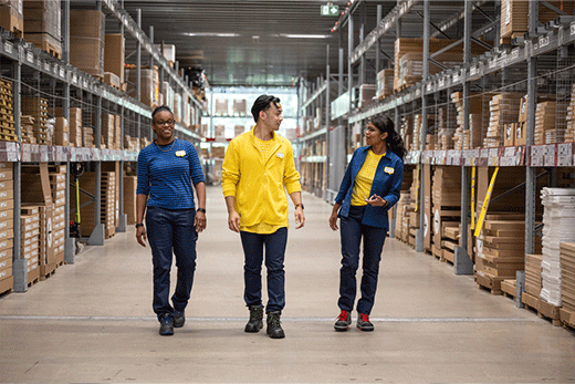 Tres colaboradores de IKEA con uniformes azules y amarillos caminan y conversan juntos por un pasillo ancho de un almacén de muebles de autoservicio.