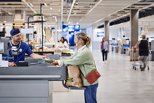 Un cajero de IKEA sonriente con uniforme de rayas azules atiende a una clienta que empaca artículos en una bolsa de papel en la línea de cajas de la tienda.