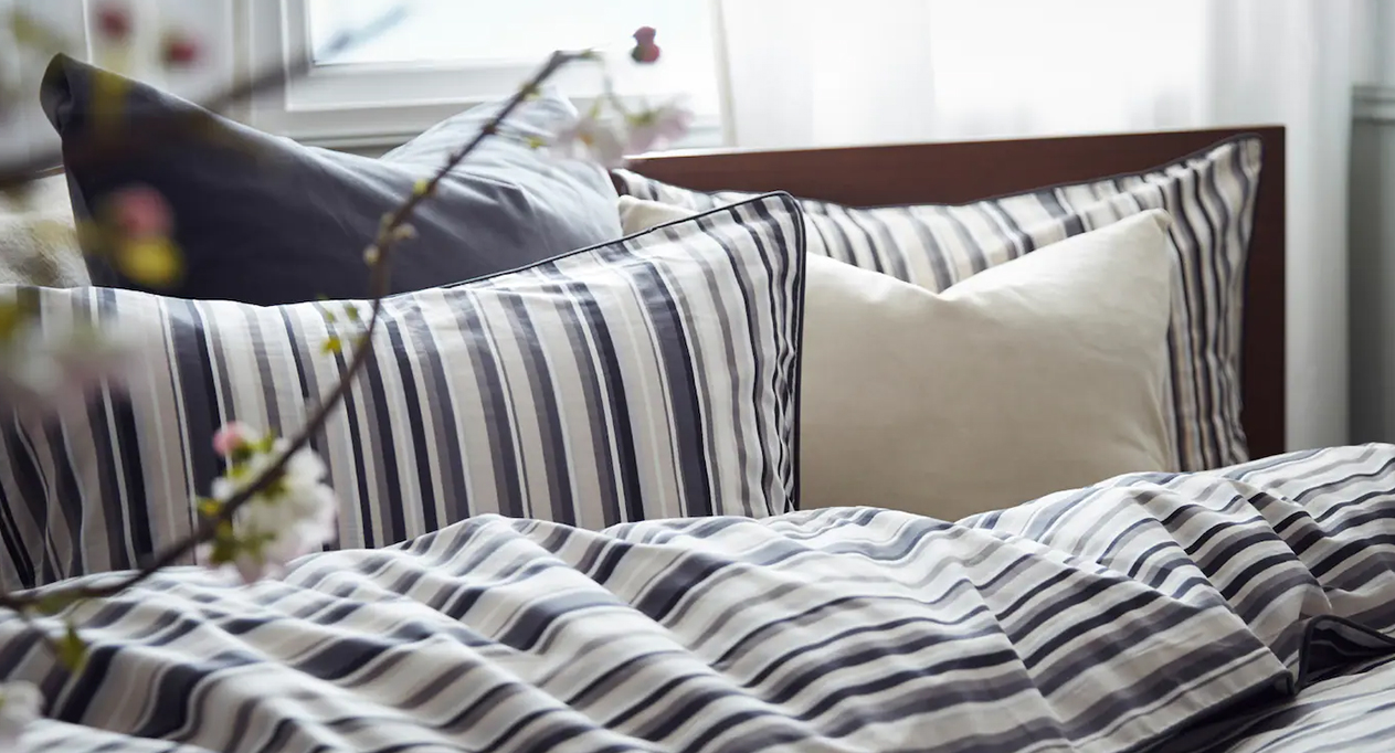 Cozy bed with striped bedding, soft pillows, and blossoms near a sunlit window create a peaceful atmosphere.