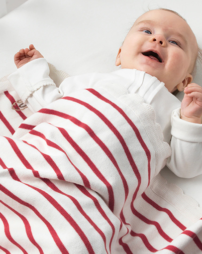 Smiling baby in white outfit, wrapped in a red-striped blanket, lying on a white bed, full of joy.