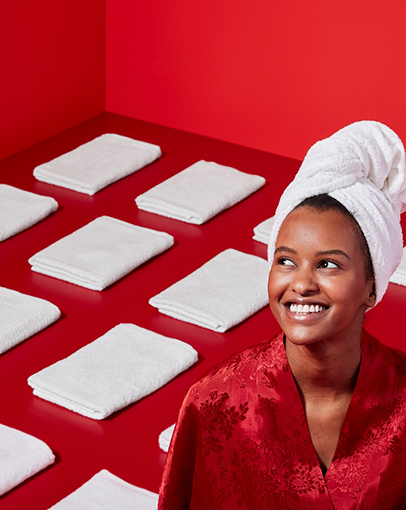 Smiling woman in red robe and towel on her head, surrounded by neatly folded white towels on a red background.