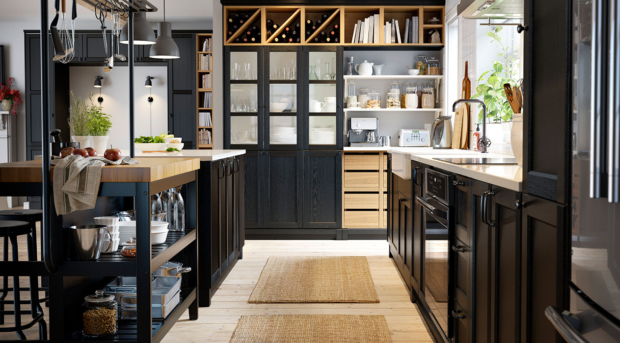 Una cocina bien iluminada con una isla VADHOLMA, a la derecha un gabinete de cocina con una placa de inducción LAGAN, y al fondo una estantería de cocina con puertas y un botellero VADHOLMA.