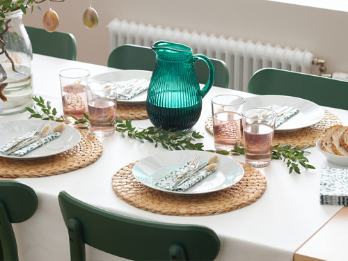 Mesa puesta con jarra verde, vajilla blanca sobre manteles individuales de fibra y sillas verdes.
