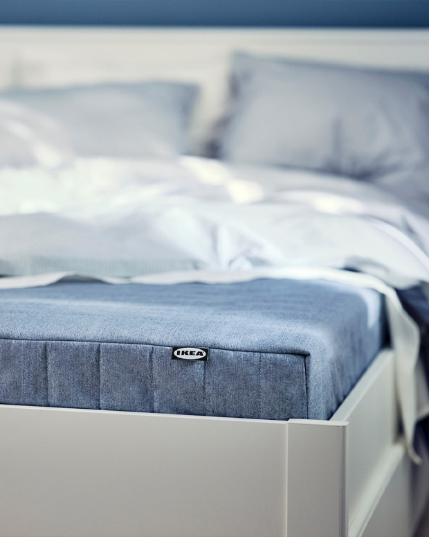 Esquina de una cama blanca de estilo moderno, con el colchón visible en azul y una colcha blanca encima.