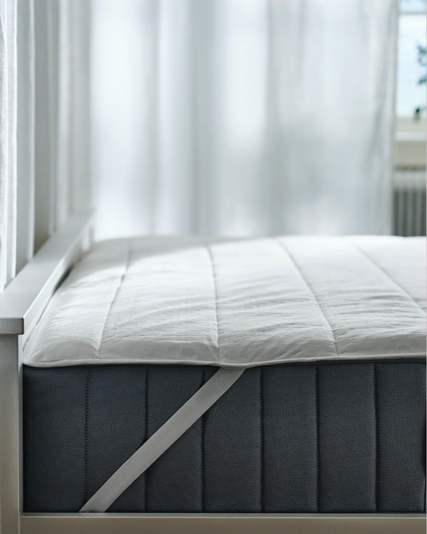Imagen de una cama con un colchón gris visible y el protector de colchón con la cinta que lo sujeta.