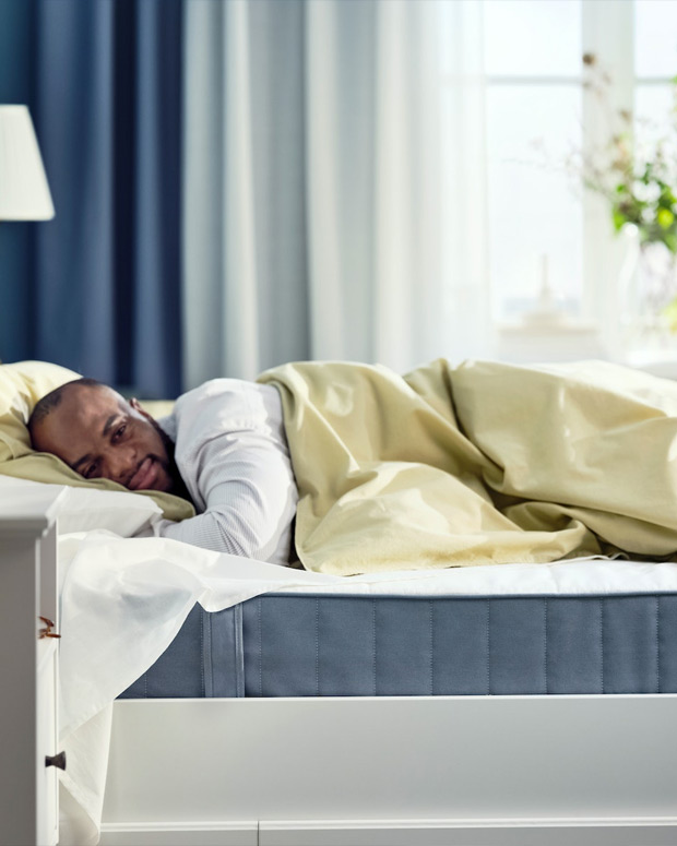 Una persona durmiendo cómodamente en una cama de estilo moderno, se ve el colchón azul, al fondo la cortina azul y blanca está desenfocada.