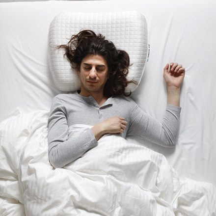 Persona tumbada boca arriba, sobre edredones blancos y con una almohada ortopédica.