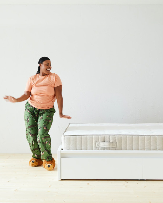 Mujer de pie en una pose dinámica delante de una cama con un colchón blanco visible, el plano está totalmente iluminado y blanco.
