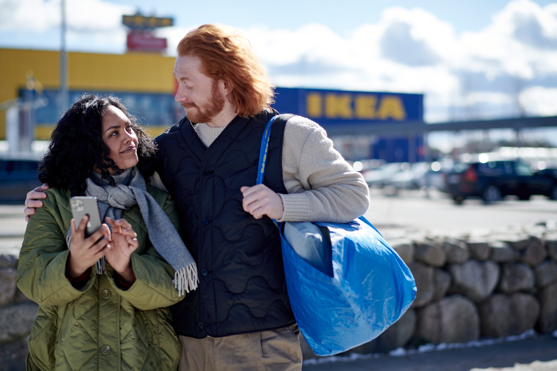 Man and woman posing together in front of the entrance of an Ikea store, with shopping bags in their hands.