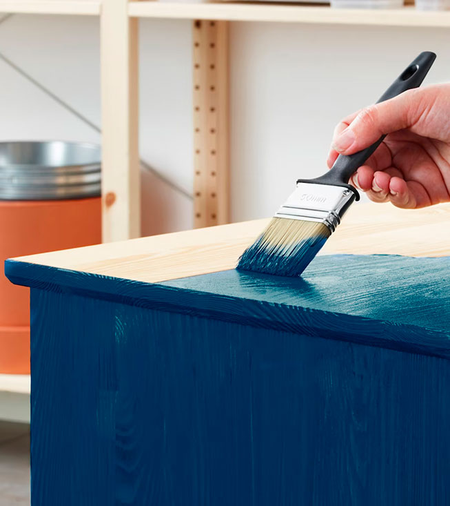 Una mano pinta una superficie de madera de color azul oscuro con una brocha, mostrando el antes y después.