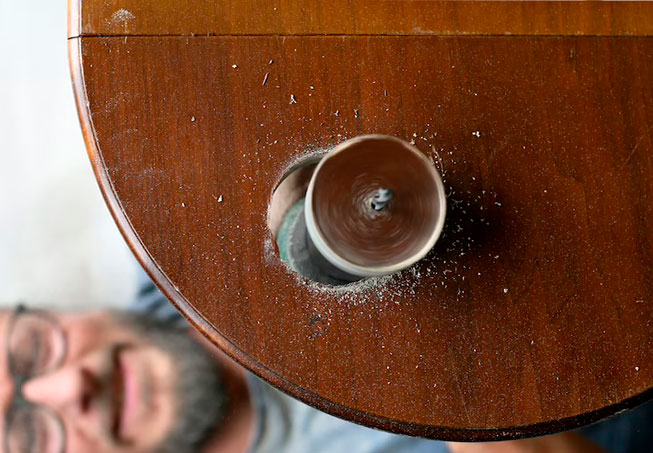 Una broca perforando una superficie de madera, con virutas. Cara borrosa de un hombre en la esquina inferior izquierda.