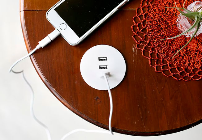 Un teléfono blanco cargándose desde un concentrador USB redondo en una mesa de madera. Tapete naranja y planta.