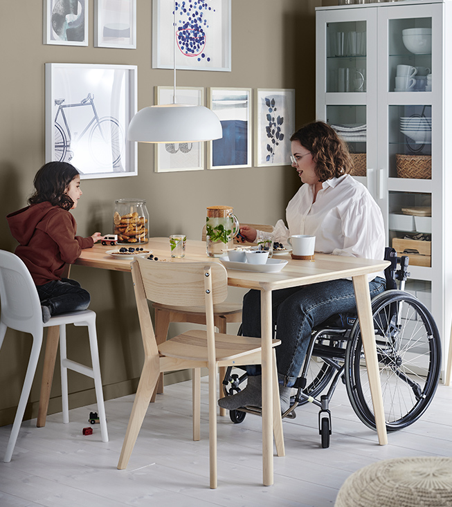  Aquí tienes la descripción en 120 caracteres para ambos idiomas:  Español:  Una mujer en silla de ruedas y un niño sentados en una mesa de comedor de madera clara en una sala luminosa.