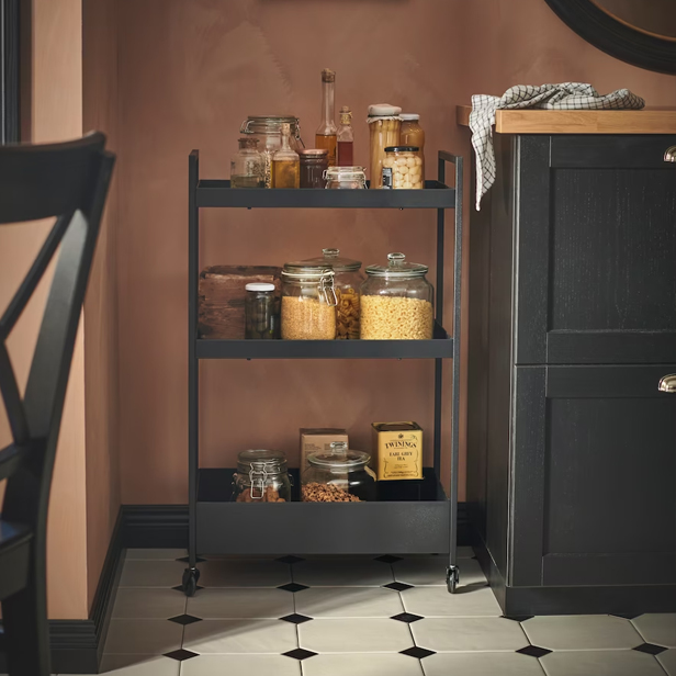 Un carrito auxiliar negro de tres niveles con frascos de comida, junto a un mueble oscuro y pared rosa.