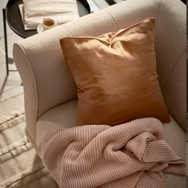 Un sofá individual beige con un cojín marrón y una manta rosa tejida, con una mesa auxiliar al fondo.
