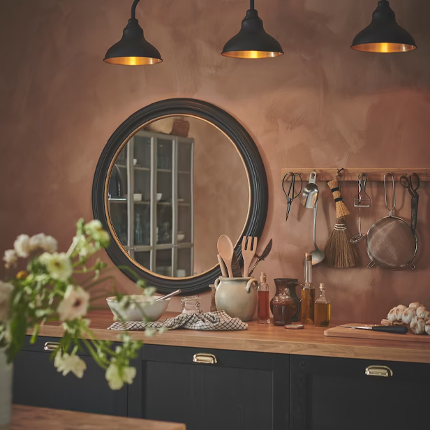 Una cocina rústica con pared rosa, espejo redondo, gabinetes negros y encimera de madera.