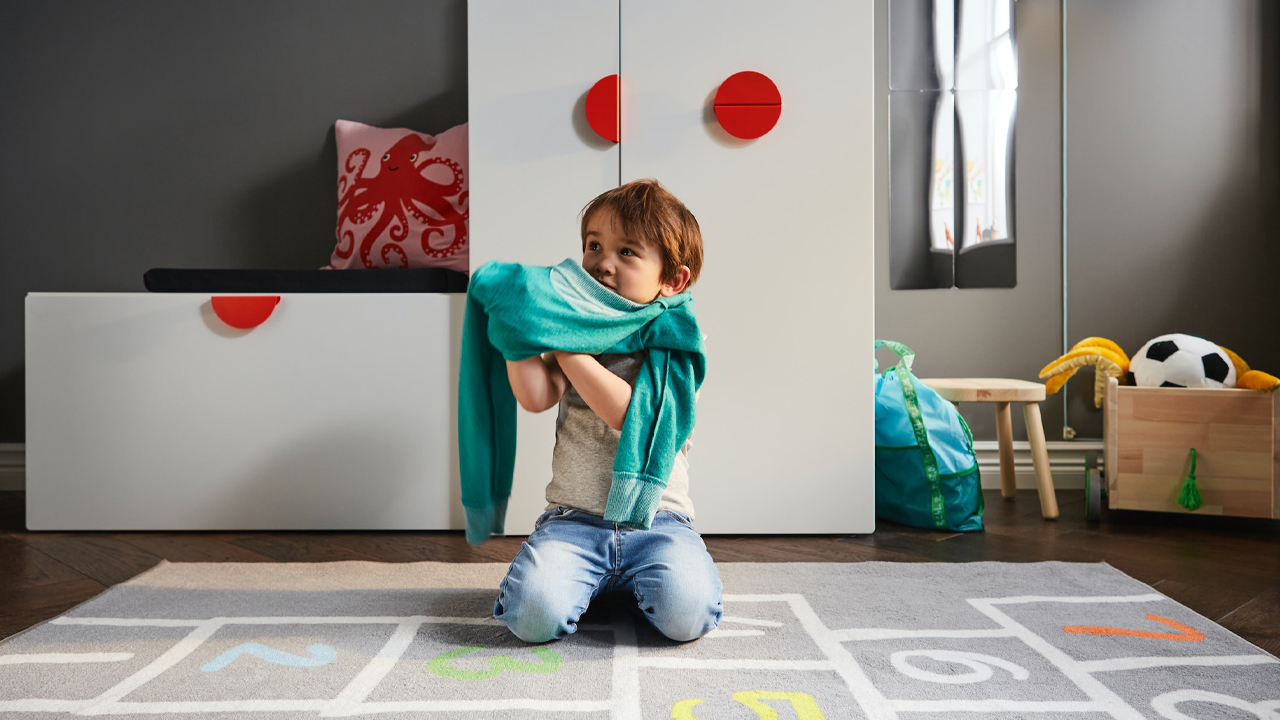 A child sitting down putting on a sweatshirt, and behind him, two SMASTAD storage units with BEGRIPA handles; to the right, a BLÅVINGAD backpack.