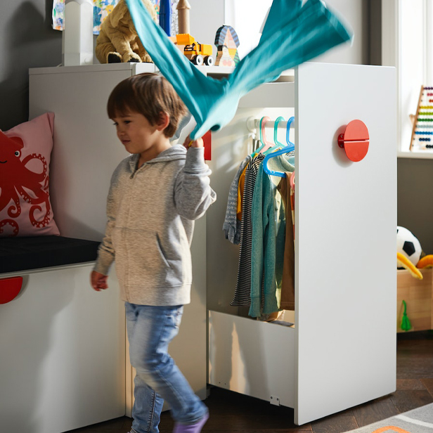 A child holding a sweatshirt with a SMÅSTAD wardrobe behind him, and a BLÅVINGAD cushion to the left.