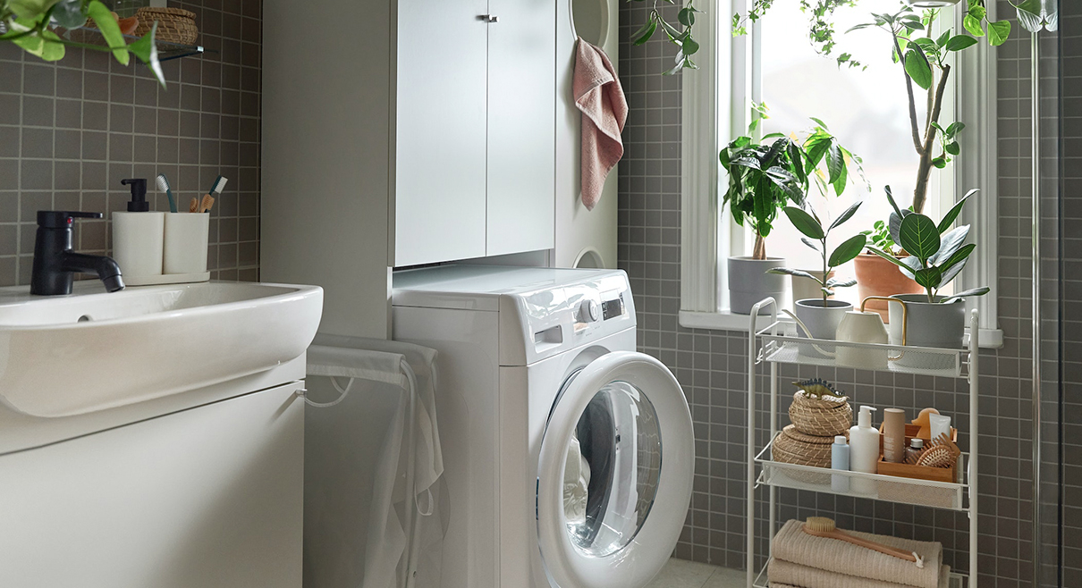 A functional bathroom with gray tiles, washing machine, sink, and plants by a window. Tidy and bright atmosphere.