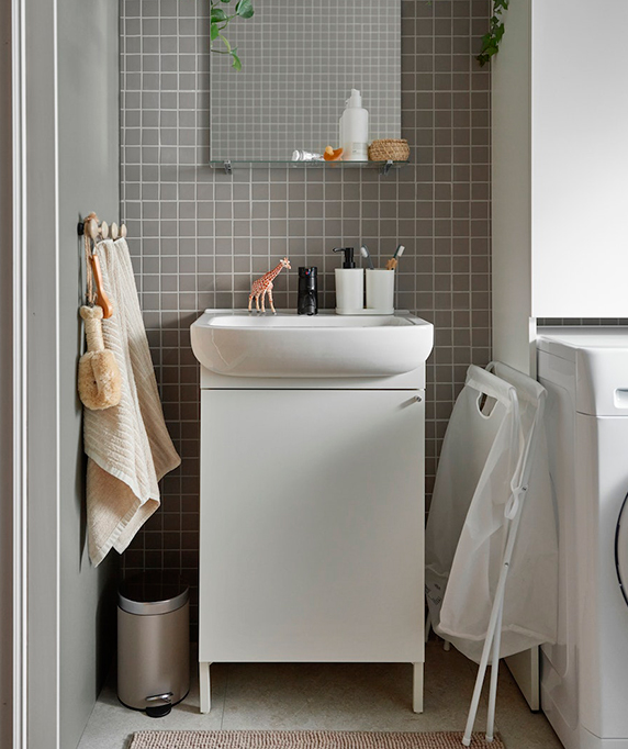 A small bathroom with gray tiles, a white sink, hanging towels, and a laundry basket. Functional and compact atmosphere.
