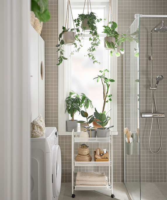 A bright bathroom with gray tiles, hanging plants, and a cart with towels near the shower. Fresh, natural ambiance.