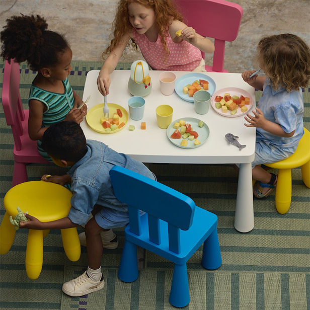 Niños sentados alrededor de una pequeña mesa blanca con sillas de colores pastel, servido platos de colores pastel servido hay frutas cuadradas