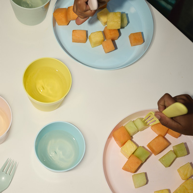 Platos de colores pastel, con fruta cortada en cuadrados, con manos de niños comiendo.
