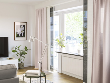 Salón luminoso con grandes ventanas, cortinas claras y lámpara de pie metálica.