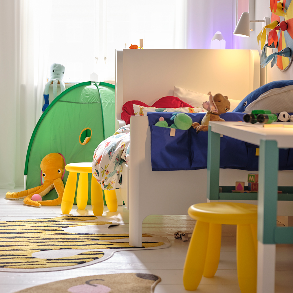 Un cuarto infantil con un sistema de almacenamiento TROFAST a la izquierda, un escritorio PAHL a la derecha con dos taburetes amarillos MAMMUT y al fondo una tienda DVÄRGMÅS.