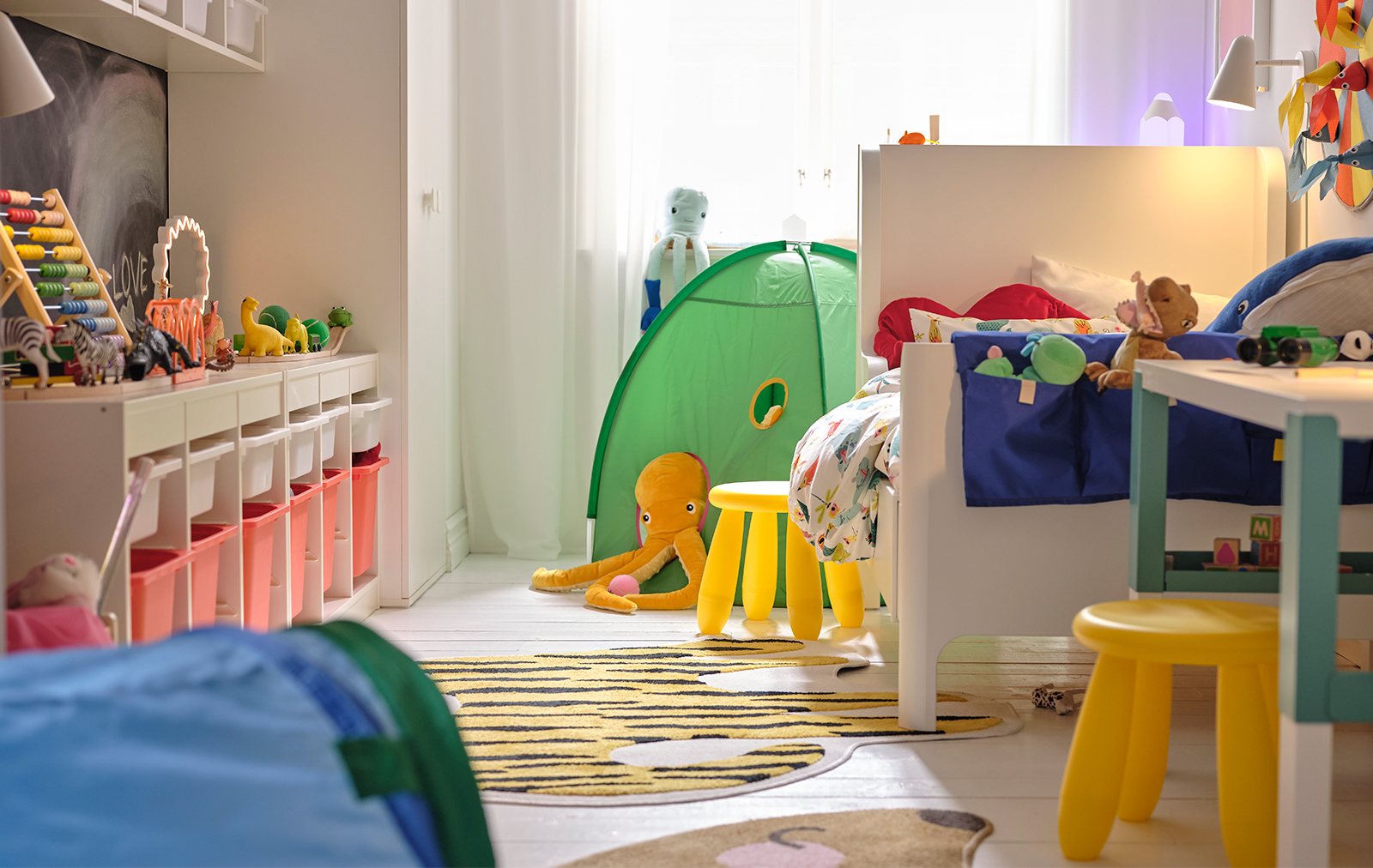 Un cuarto infantil con un sistema de almacenamiento TROFAST a la izquierda, un escritorio PAHL a la derecha con dos taburetes amarillos MAMMUT y al fondo una tienda DVÄRGMÅS.