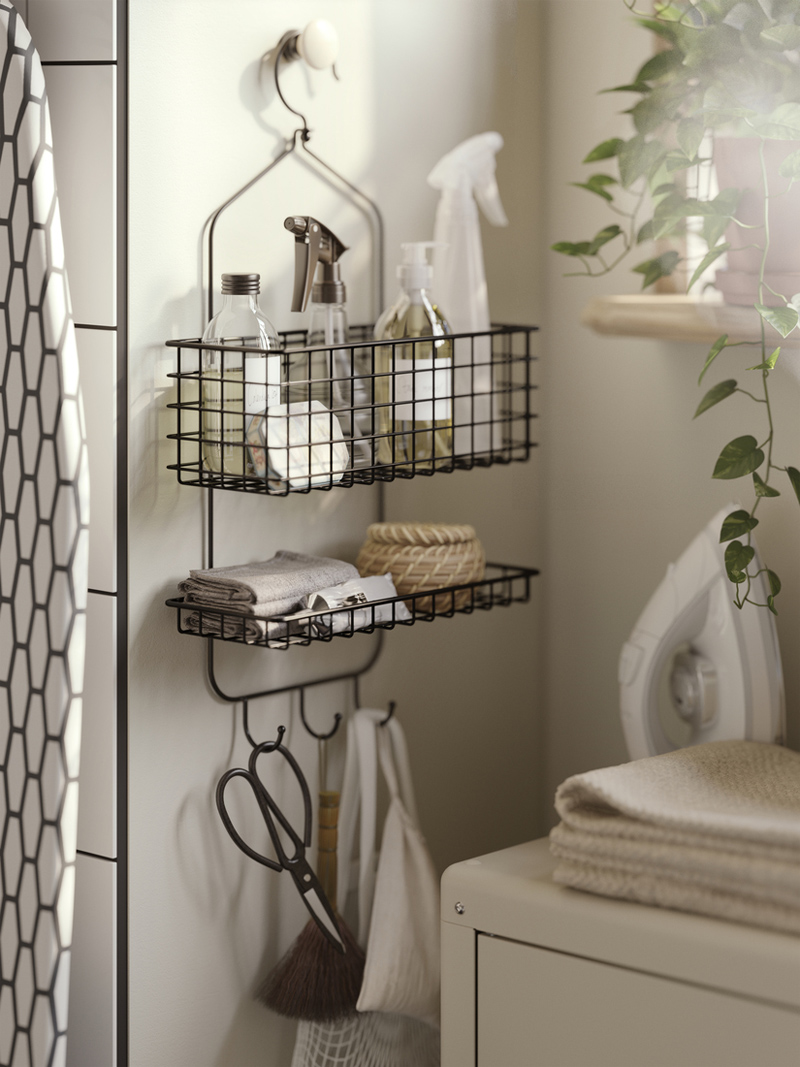 Organizador de pared BLECKSJÖN de alambre negro con botellas de limpieza, toallas y cesta bajo luz suave.
