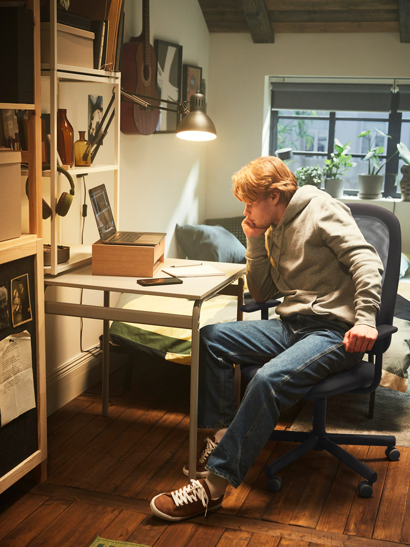 Red-haired man working on laptop at a desk with shelving, warm light, and a bohemian atmosphere.