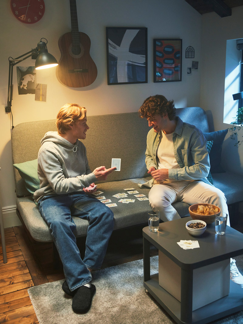 Two friends play cards on a gray sofa, with a relaxed atmosphere and musical and artistic touches.
