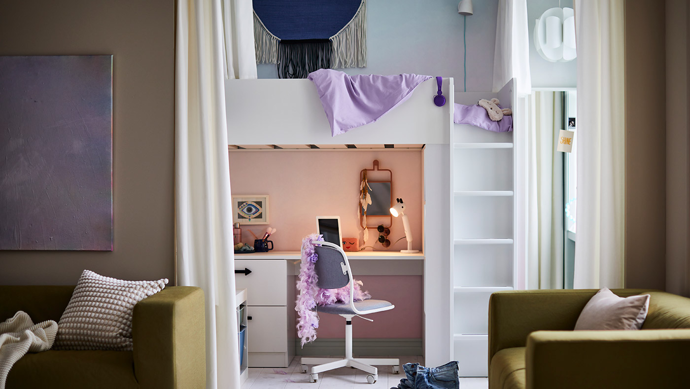 En la imagen se observa una habitación juvenil con una cama alta blanca, un escritorio debajo con silla, sofás verdes y decoración lila. El ambiente es moderno y funcional.