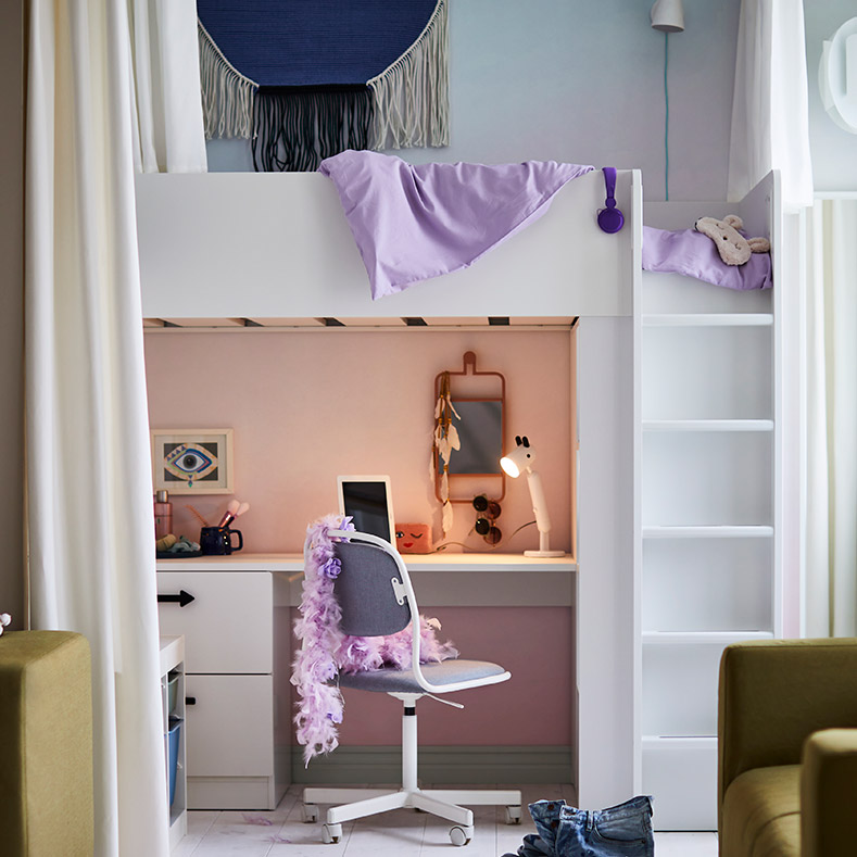 En la imagen se observa una habitación juvenil con una cama alta blanca, un escritorio debajo con silla, sofás verdes y decoración lila. El ambiente es moderno y funcional.