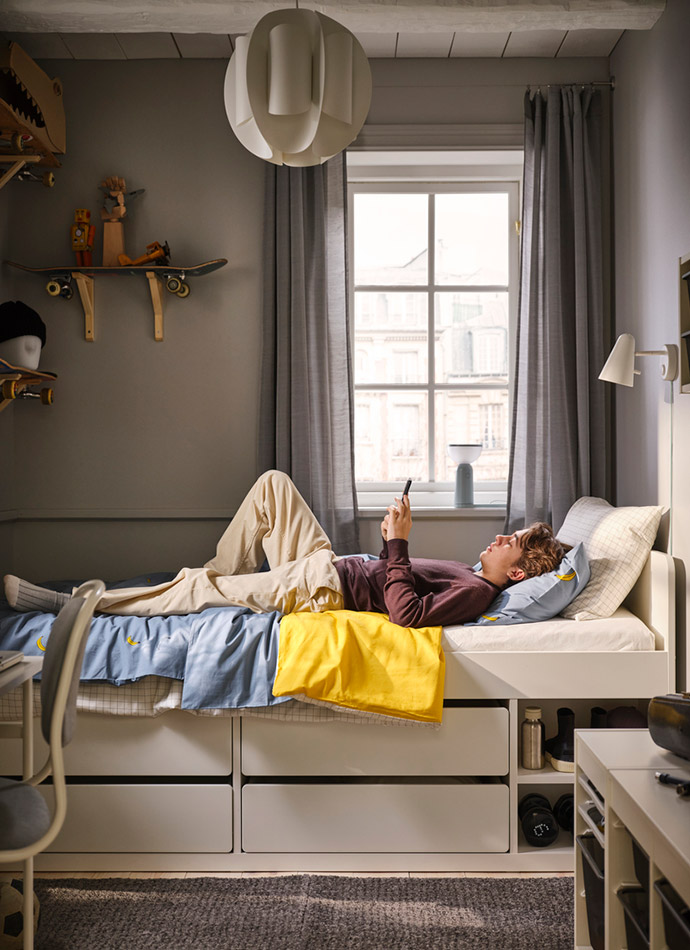 Una habitación juvenil con paredes grises, una cama con cajones, sábanas azules y una manta amarilla. Un joven usa su teléfono.