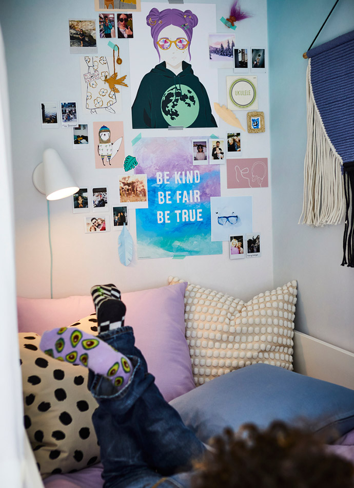 En la imagen se ve una pared con pósters coloridos y fotos, cojines de tonos lilas y beige, y calcetines divertidos. Ambiente juvenil y personalizado.