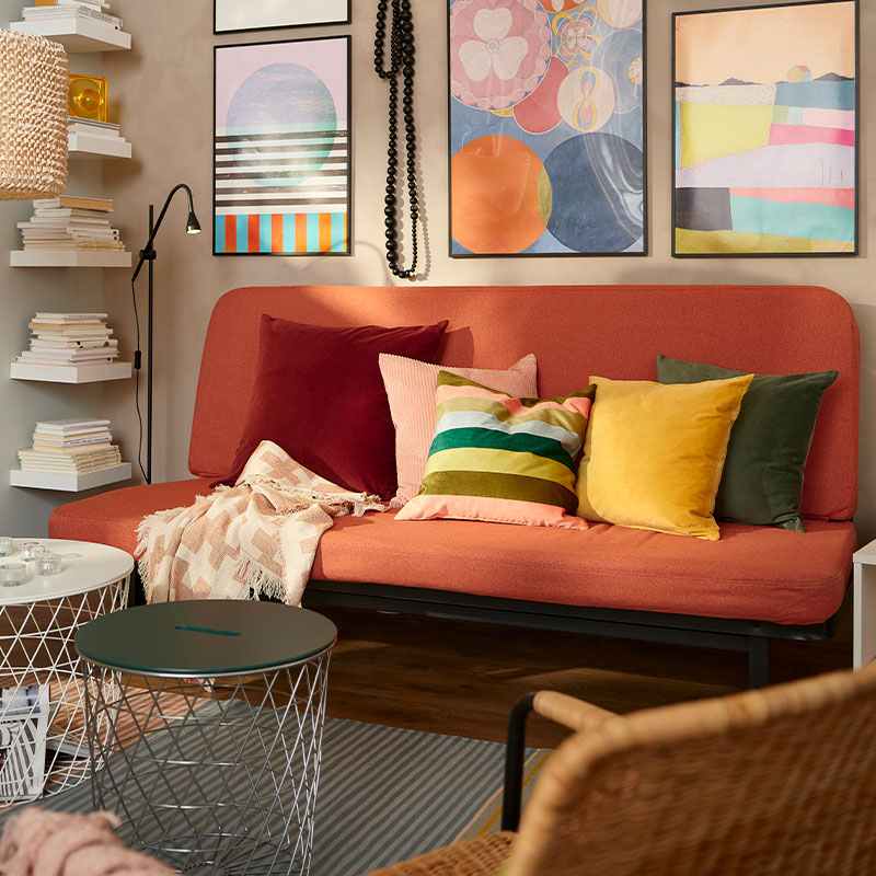 Ambiente colorido con un sofá minimalista con varios cojines multicolores, estilo años 70, hay una estantería abierta de madera de pino,
