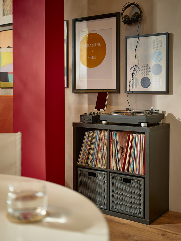 armario abierto con cuatro espacios, con cajas de mimbre en el inferior y lleno de discos de vinilo en la parte superior, hay un tocadiscos en la parte superior, y en la pared fotos de discos.