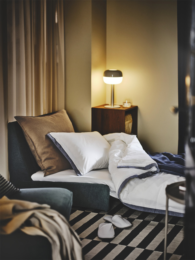 Elegant and modern corner with single sofa bed, duvet cover with cushions, neutral-toned curtains, and nightstand with lit lamp