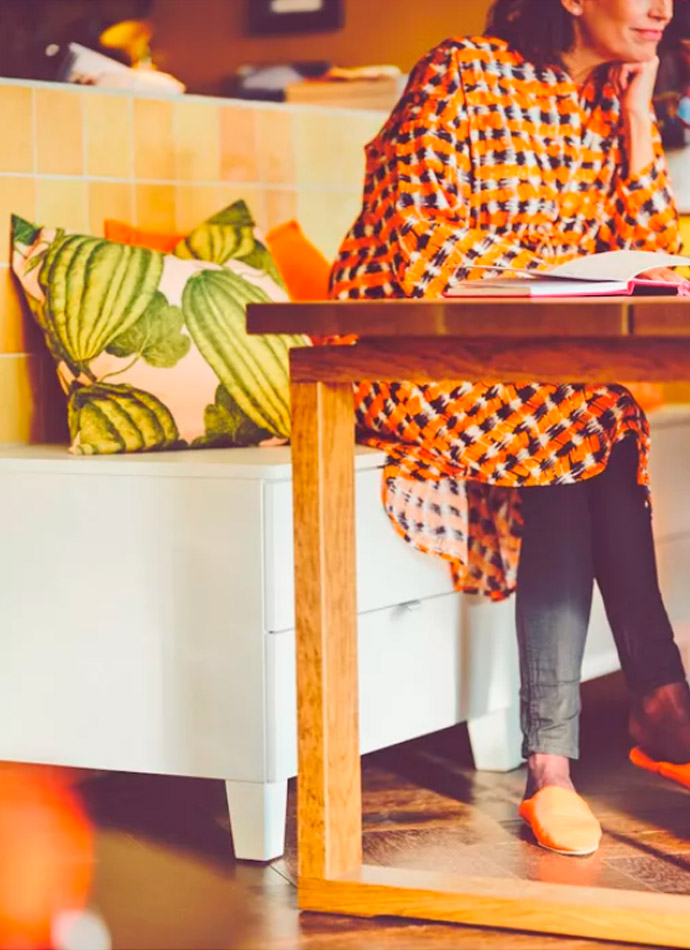 A woman in an orange and black tunic sits on a white bench with vibrant cushions. Warm, casual atmosphere.