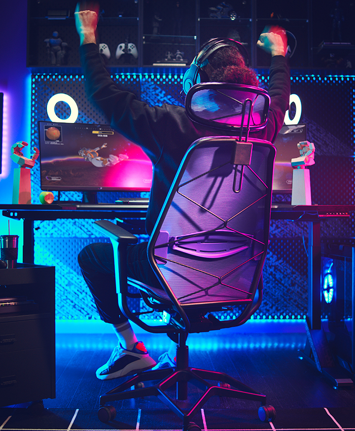 Un jugador celebra victoria en cuarto iluminado por luces neón azul y rosa. Sentado en silla de malla negra frente a PC con monitor "O" brillante.